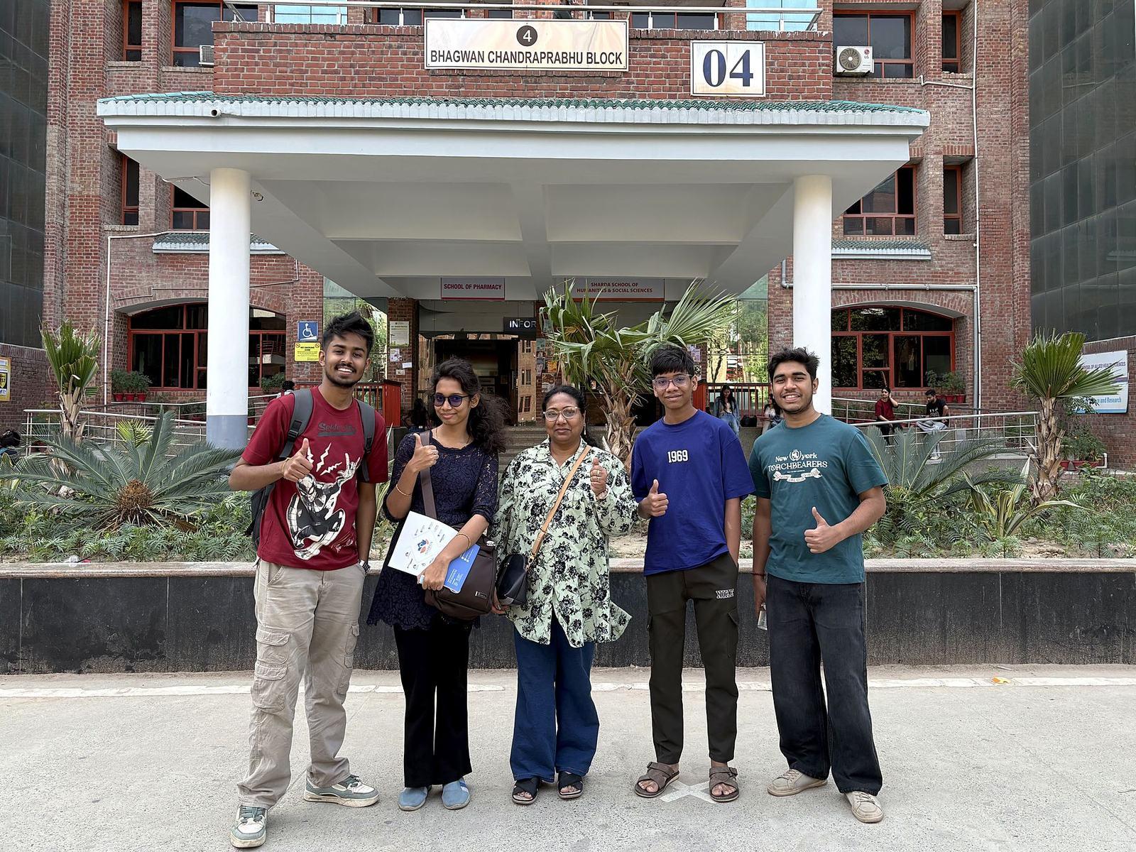 Sharda University ক্যাম্পাসে ব্লক ০৪ (Bhagwan Chandraprabhu Block)-এর সামনে পাঁচজন হাসিমুখে থাম্বস-আপ দিচ্ছেন, পেছনে লাল ইটের ভবন